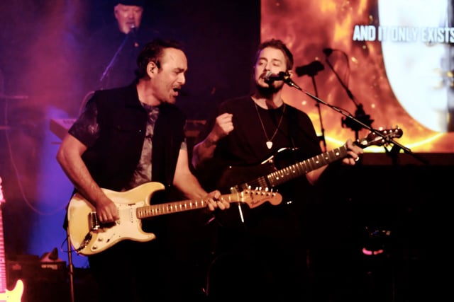Neal Morse and a guitarist singing side by side, both playing electric guitars under warm stage light