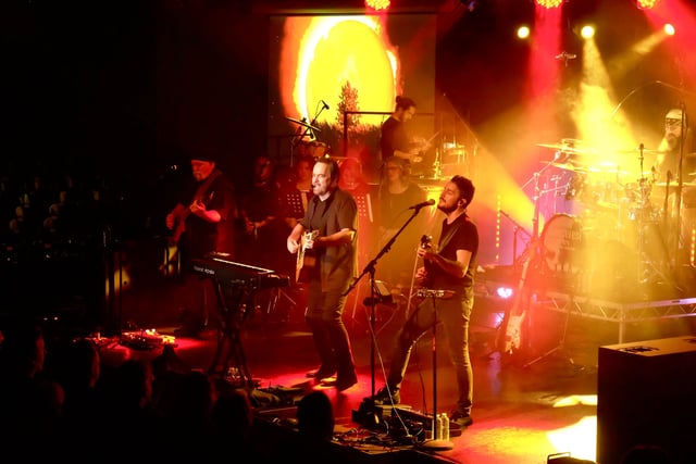 Neal Morse playing acoustic guitar beside a guitarist, the band behind them under a glowing orange sun on the screen