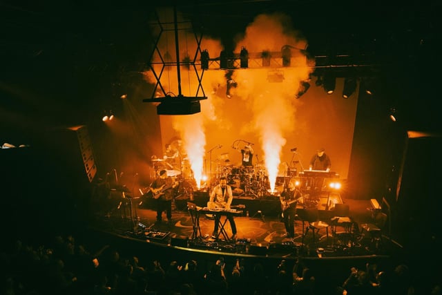 The band on a theater stage seen from the balcony, with columns of smoke lit orange rising behind them